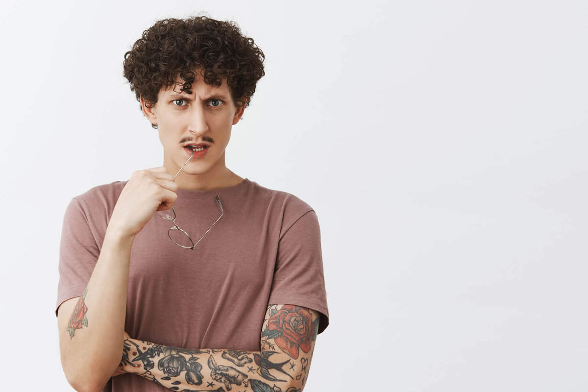Male with tattooes curly hair and moustache biting rim of glasses and looking to ask a question about laser tattoo removal
