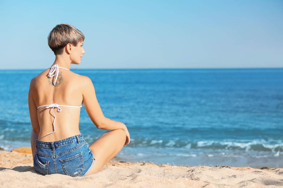 Woman with back tattoo sitting on the beach looking out to sea
