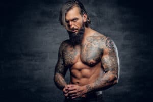 Tanned man with long hair and beard topless with a full sleeve and chest tattoo to reflect tattoo trends in the 1950s
