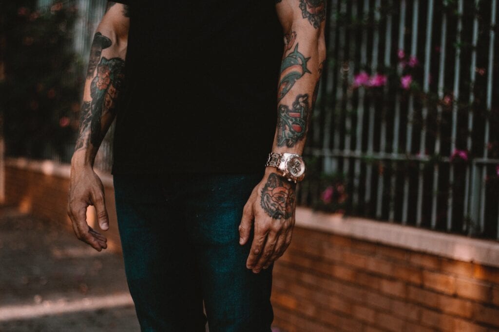 A tattooed man wearing a black shirt, standing near a slatted fence, captured in an outdoor setting.