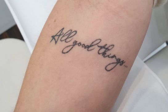 Showing cursive “All good things” tattoo script inked on a forearm with clean black linework.