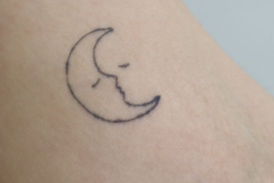 Showing small crescent moon face tattoo inked on an arm with fine black outline and minimalist styling.