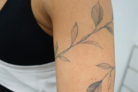 Showing fine line botanical arm tattoo with delicate leafy branches wrapped around the upper arm.