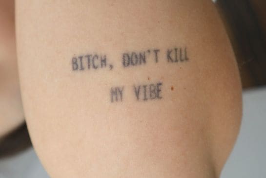 Showing small quote tattoo on the tricep with faded black text inked in a minimalist typewriter style.
